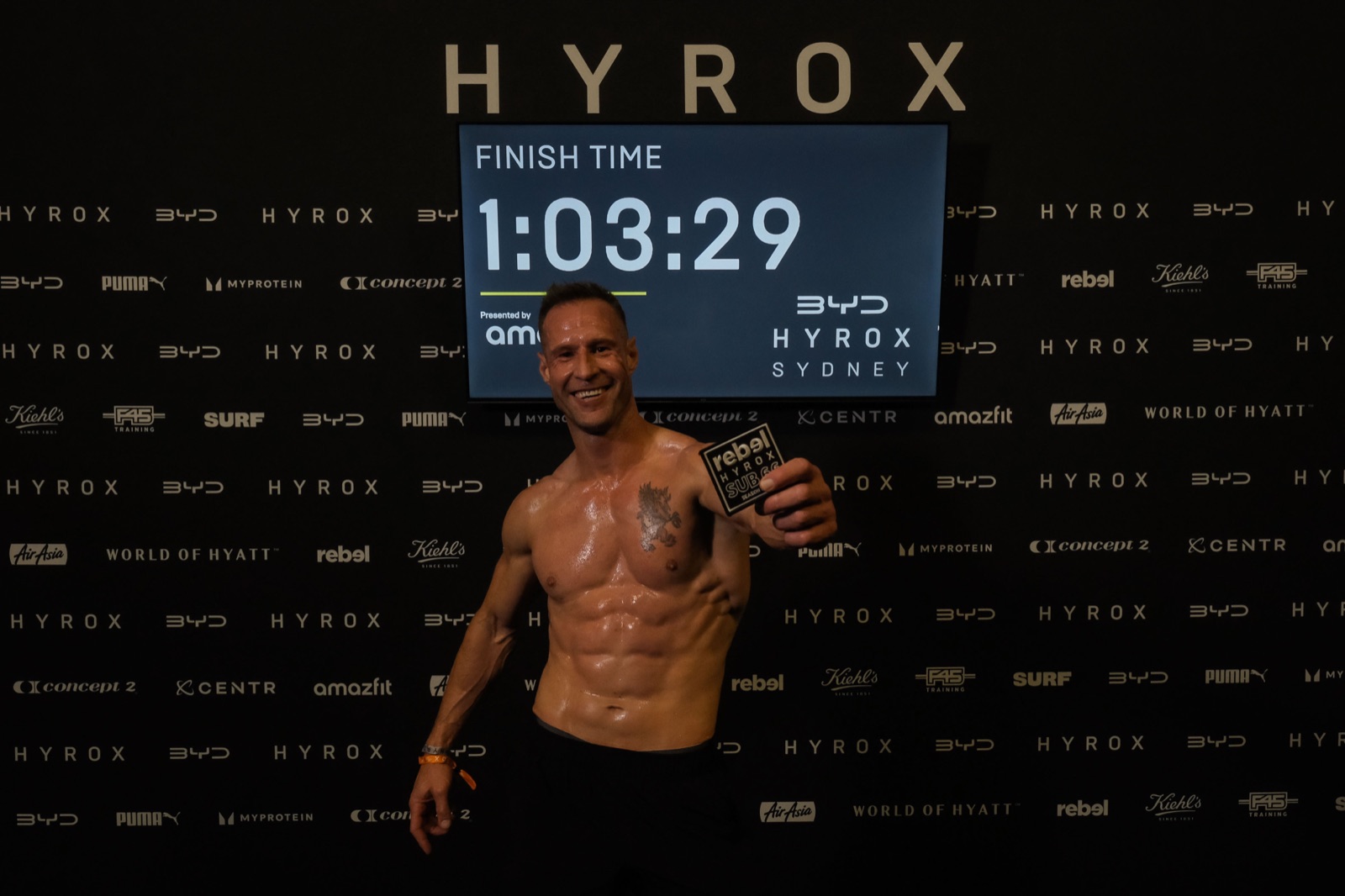 Tyson Schubert holding his HYROX finisher patch in front of a screen showing a 1 hour 3 minute 29 second finish